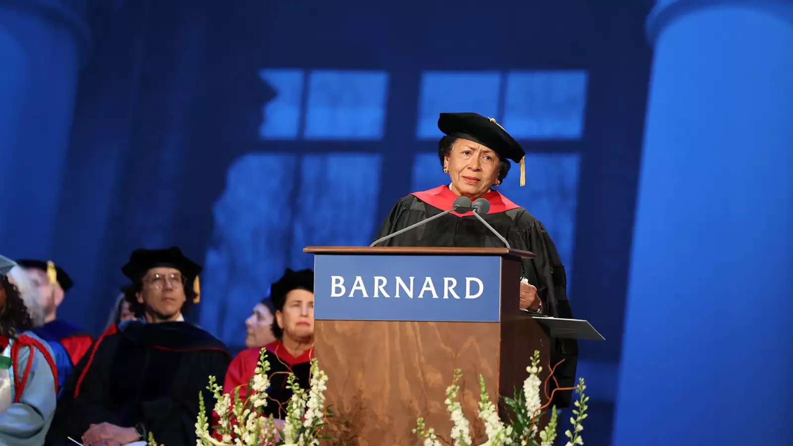 Older woman dressed in college regalia at lecturn