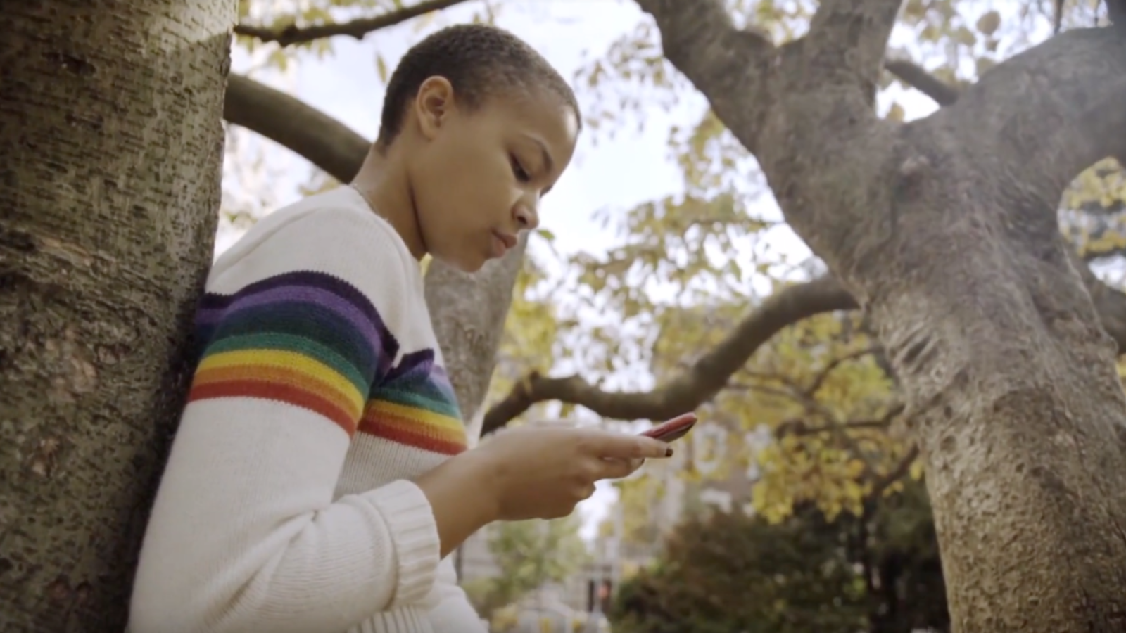 Black woman student leaning against a tree, looking at her phone