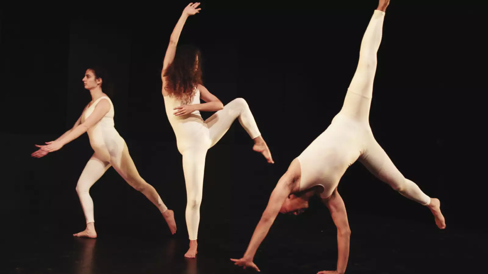 3 people in white body suits dancing on a black stage