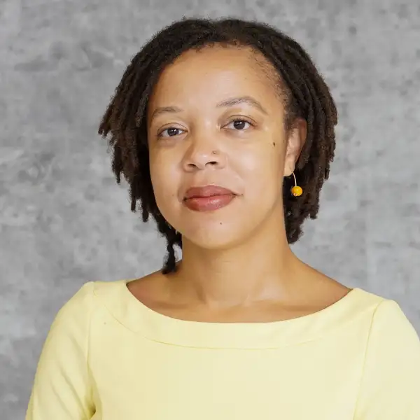 Headshot image of Dr. Amelia Simone Herbert. She has short locs and a yellow dress and looks directly into the camera.