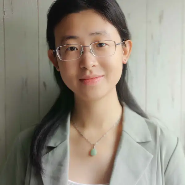 Professor Xia smiles gently at the camera. She is wearing a pale green-gray blazer with a white shirt underneath. Her medium length hair is half up and half down. She is wearing a necklace with a silver chain and pale green stone. Behind her is wood painted pale green.