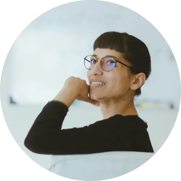 Gabrielle Gutierrez wears a black shirt, glasses, and has bangs. She smiles with her mouth closed and looks over her shoulder, resting her chin on her hand. 