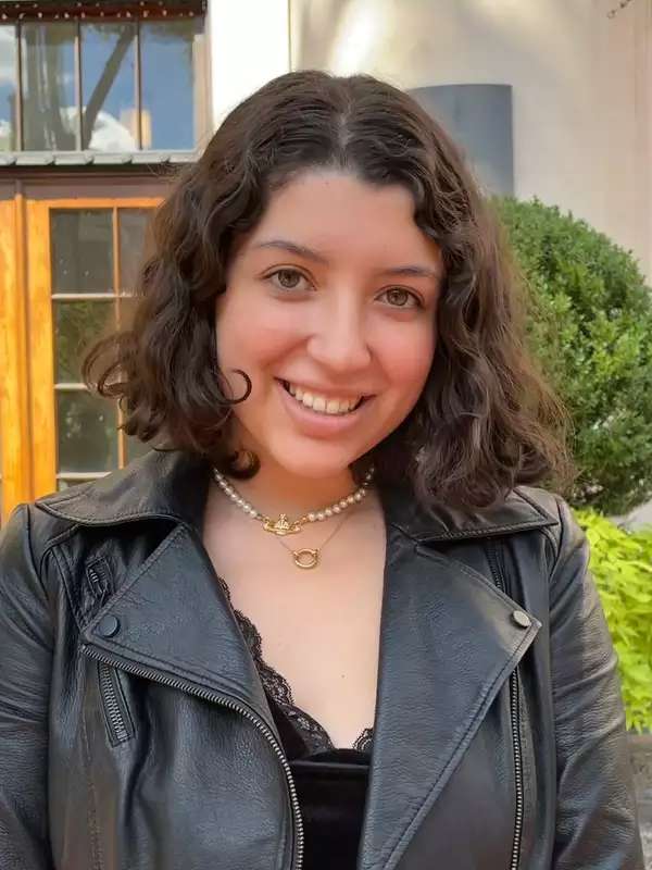 Isa stands in a black leather jacket in front of a window and some bushes and smiles at the camera.