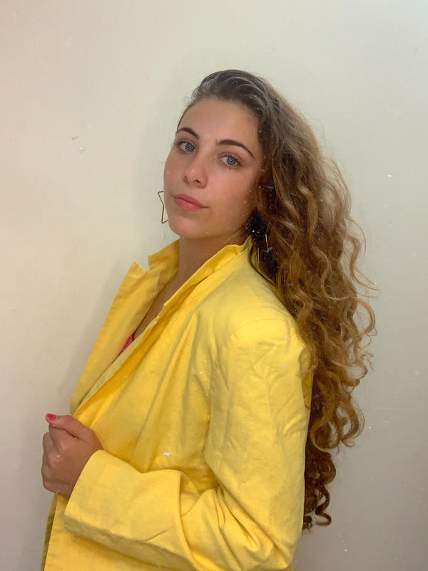 Izzy is photographed in front of a white wall, wearing a yellow jacket and star shaped earrings.