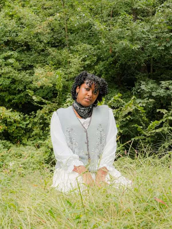 Image of Ashe kneeling in a field of grass and trees in the background, with his hands in his lap and a soft smile
