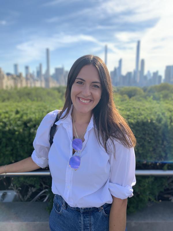 Nicole is wearing a white shirt and smiling in front of a view of the New York City skyline.