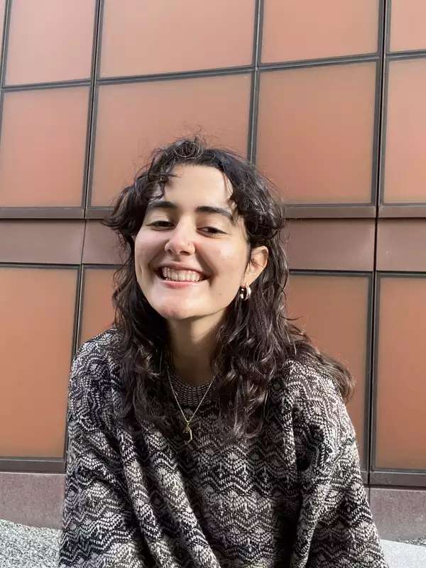 Nia (a Barnard student) is wearing a chunky brown sweater and sitting in front of an orange external wall of the Diana Center.