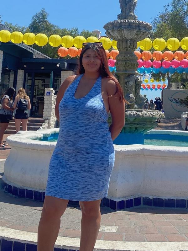 Jennifer stands in a blue dress in front of a fountain on a sunny day and is smiling at the camera. There are yellow lanterns in the background.