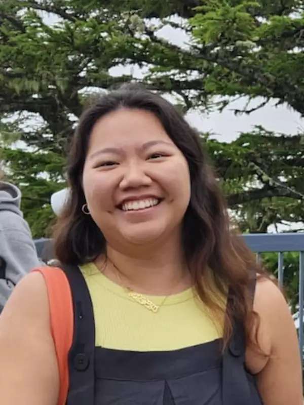 Sakira wears a yellow shirt and smiles at the camera in the foreground, with trees in the background.
