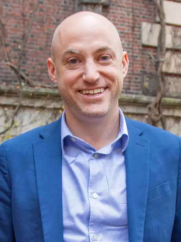 A-J Aronstein smiling in front of a brown building wearing a collared button up shirt (lavender) and a blue blazer