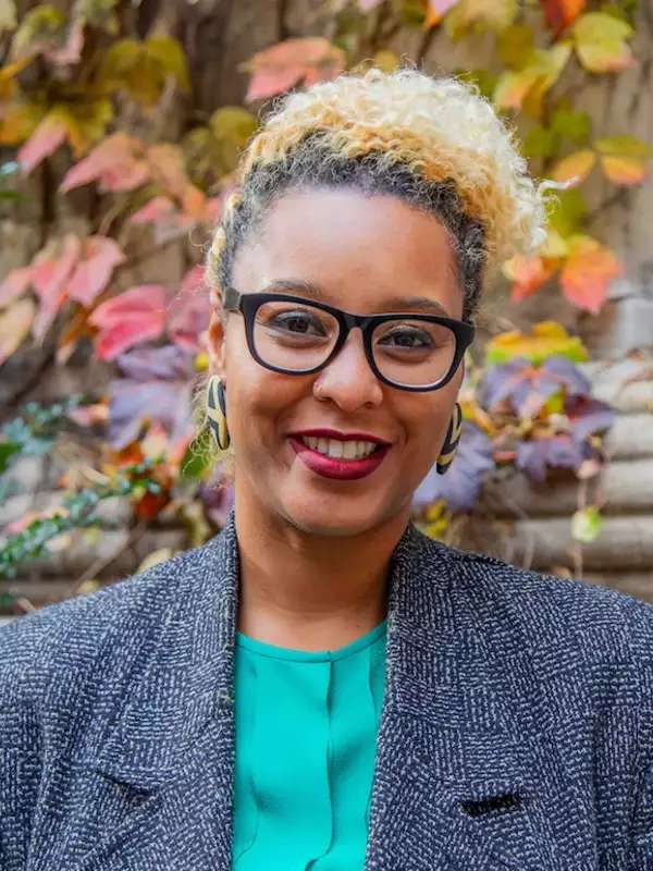 fall background, woman smiling with black and blonde hair, wearing glasses and gray coat with green top