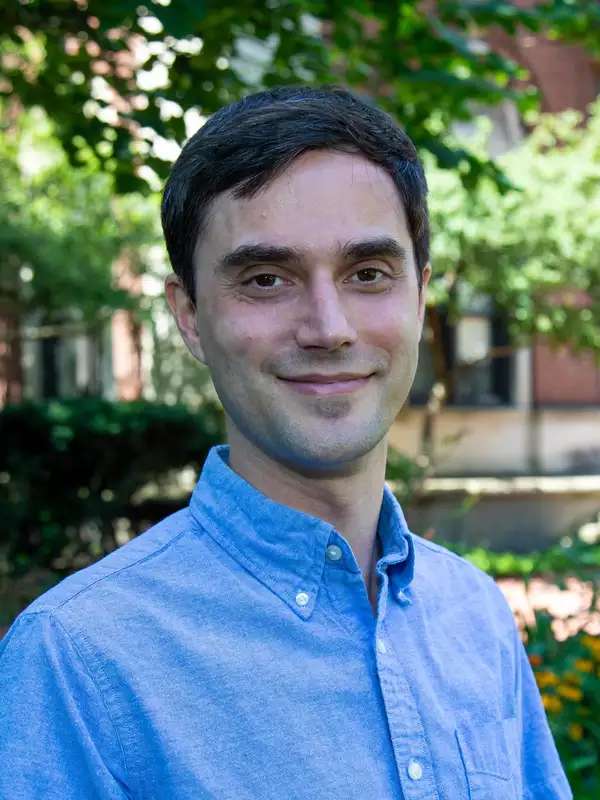 Alex Pittman smiles outside in a collared blue shirt in front of the lush background of Barnard's campus. 