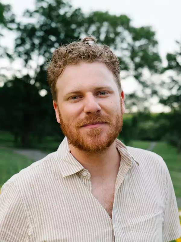 A photo of Drew Chambers. He is white and has curly auburn hair, a beard, and green eyes. He is wearing a beige and white striped shirt. There is foliage behind him.