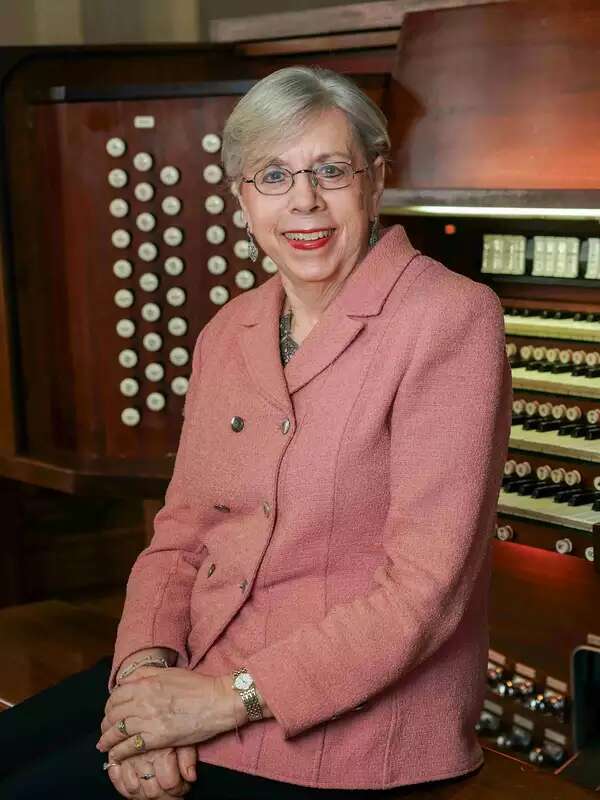 Gail Archer sits in front of an organ wearing a pink blazer. She leans on the organ and holds her hands on top of each other in front of her torso. She is smiling and looks at the camera. She wears dangling earrings, a necklace that is barely visible under the blazer, a watch and bracelets, and rings. She is a white woman who wears round rectangular wire-frame glasses. She has short grey hair that stops right above her ears. She wears eyeliner and red lipstick in the photo.