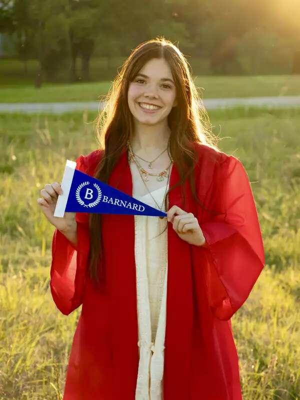 A student in a red graduation gown stands outside, holding a blue Barnard banner