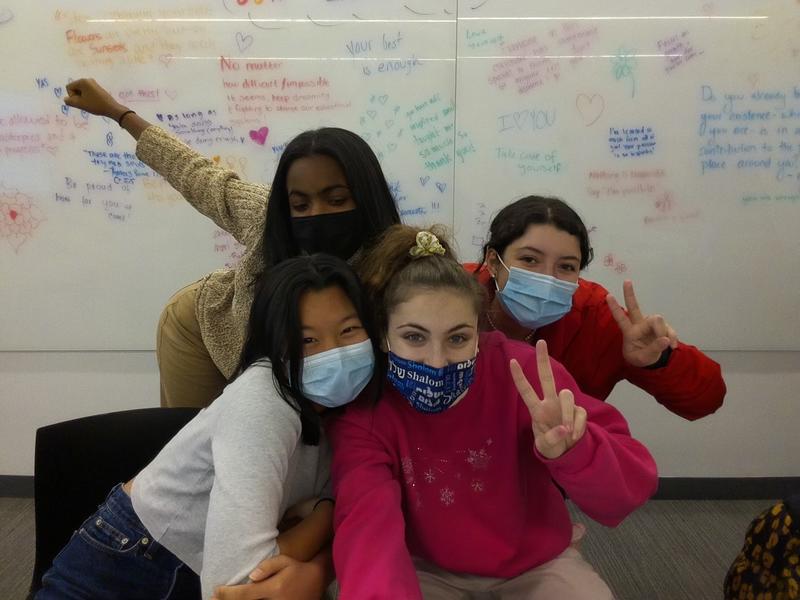 Four Barnard students who work as STEAM Fellows pose with peace signs and colorful clothing in front of a whiteboard with encouraging statements written onto it.