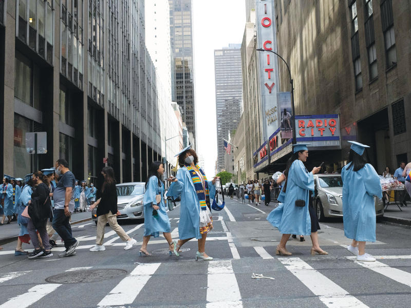 Barnard women in graduation gowns stride across a city crosswalk