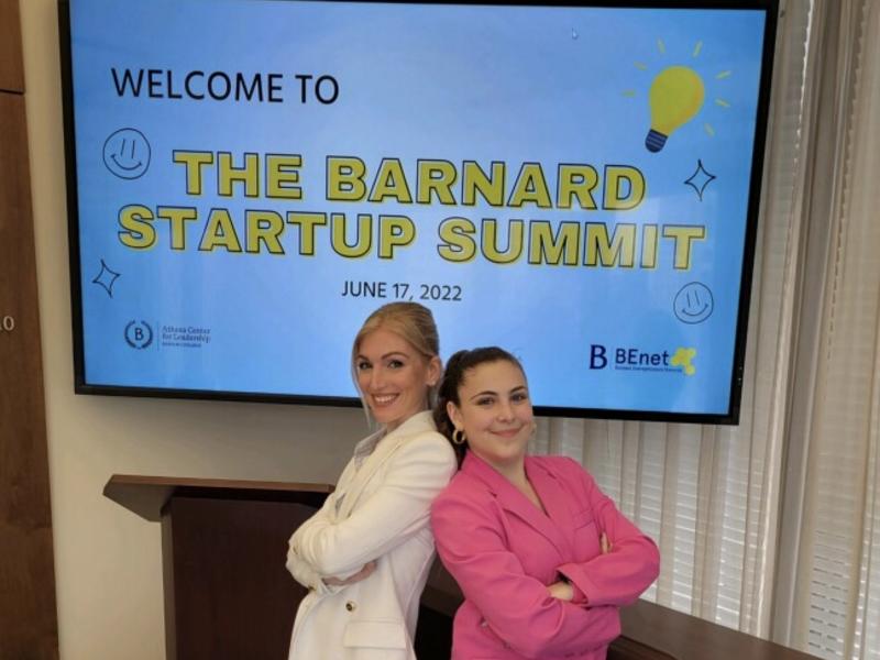 [Image description: Jessica Perusini in a white blazer stands back to back with Izzy Lapidus '24 in a pink blazer in front of a screen that says "Welcome to the Barnard Startup Summit".]