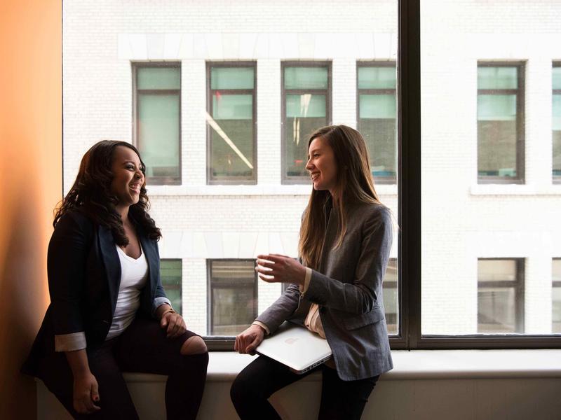 Two women in business attire chat on a window ledge.