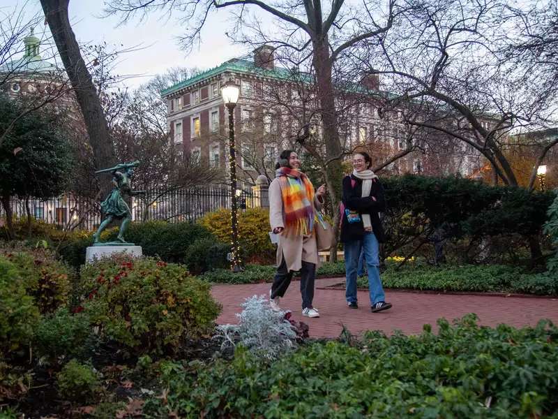 Students walk on Barnard campus with holiday lights and runner statue in background