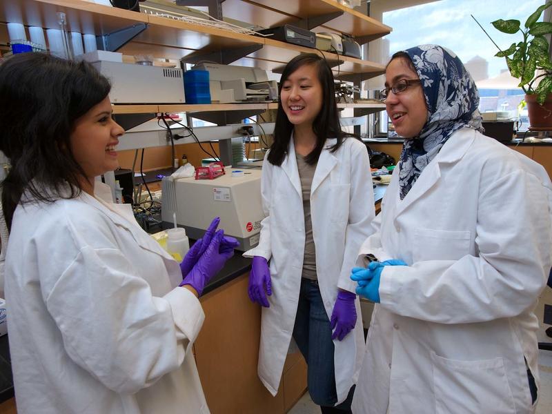 3 young women of color in white lab coats discuss something in a lab