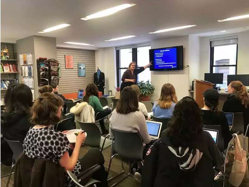 Woman pointing at screen while speaking to an audience of 12 or so students