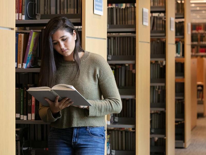 Young woman leans against a shelf of books in the library while looking at a book