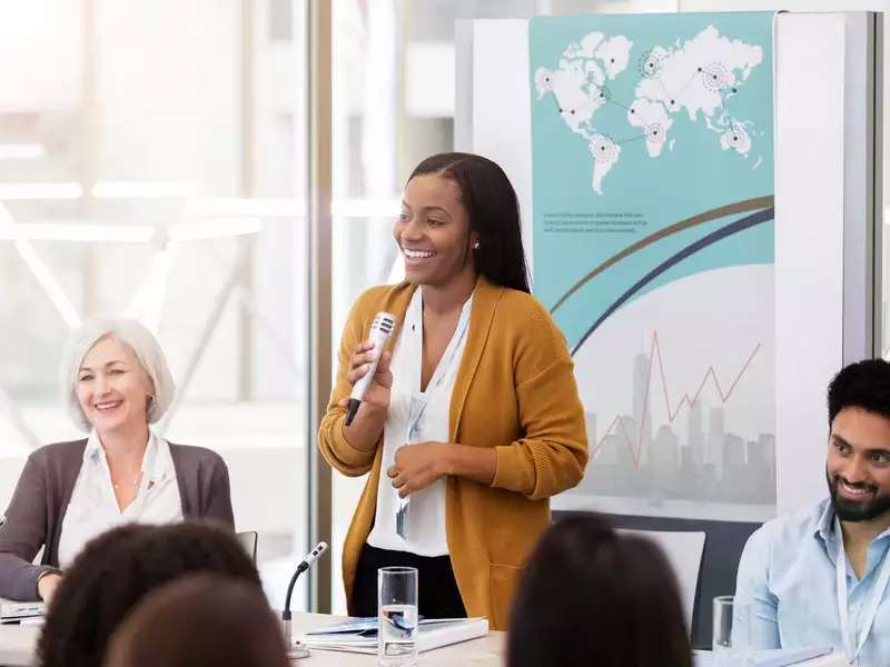 A Black woman with a microphone speaking at a panel, 2 other speakers seated