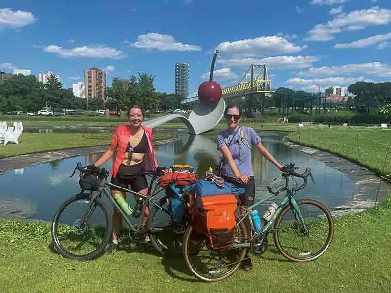 The author and her friend Brenna Forristall ’18 smile while posing with their bicycles in front of a lake with blue sky and fluffy clouds behind them