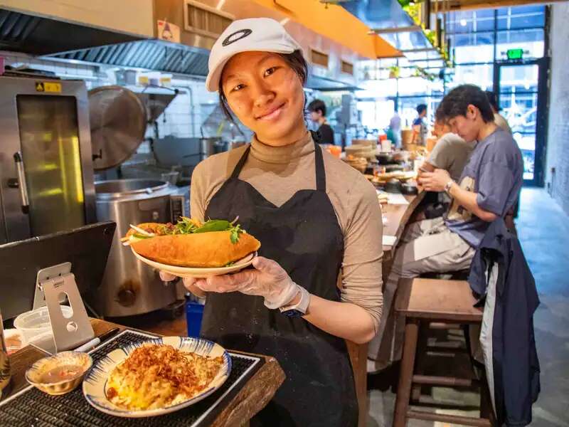Young woman wearing a cap and apron inside of a kitchen holding a bahn mi