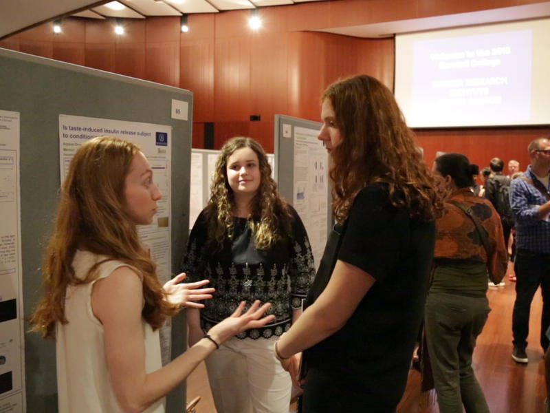 3 white women students talking in front of a large poster 