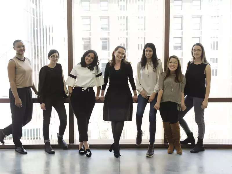 Several young women standing in a row in front of floor to ceiling windows